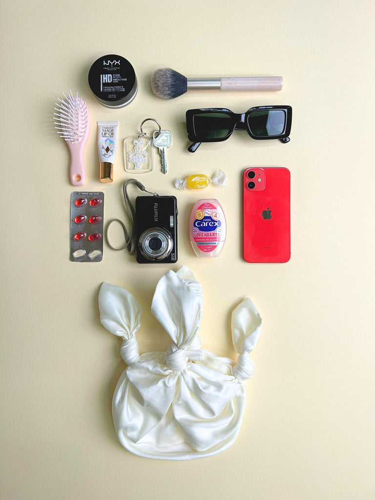 Collection of personal items including a phone, camera, makeup products, and a hairbrush on a beige background. Ivory knotted handbag sits at the bottom.