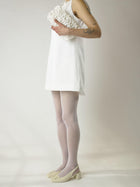 Person wearing a short ivory wedding, sheer white stocking and heeled ivory ballet pumps, holds an ivory satin textured ruched front handbag on their shoulder. Set on a white background.