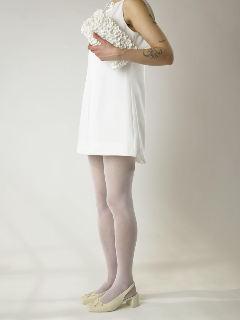 Person wearing a short ivory wedding, sheer white stocking and heeled ivory ballet pumps, holds an ivory satin textured ruched front handbag on their shoulder. Set on a white background.