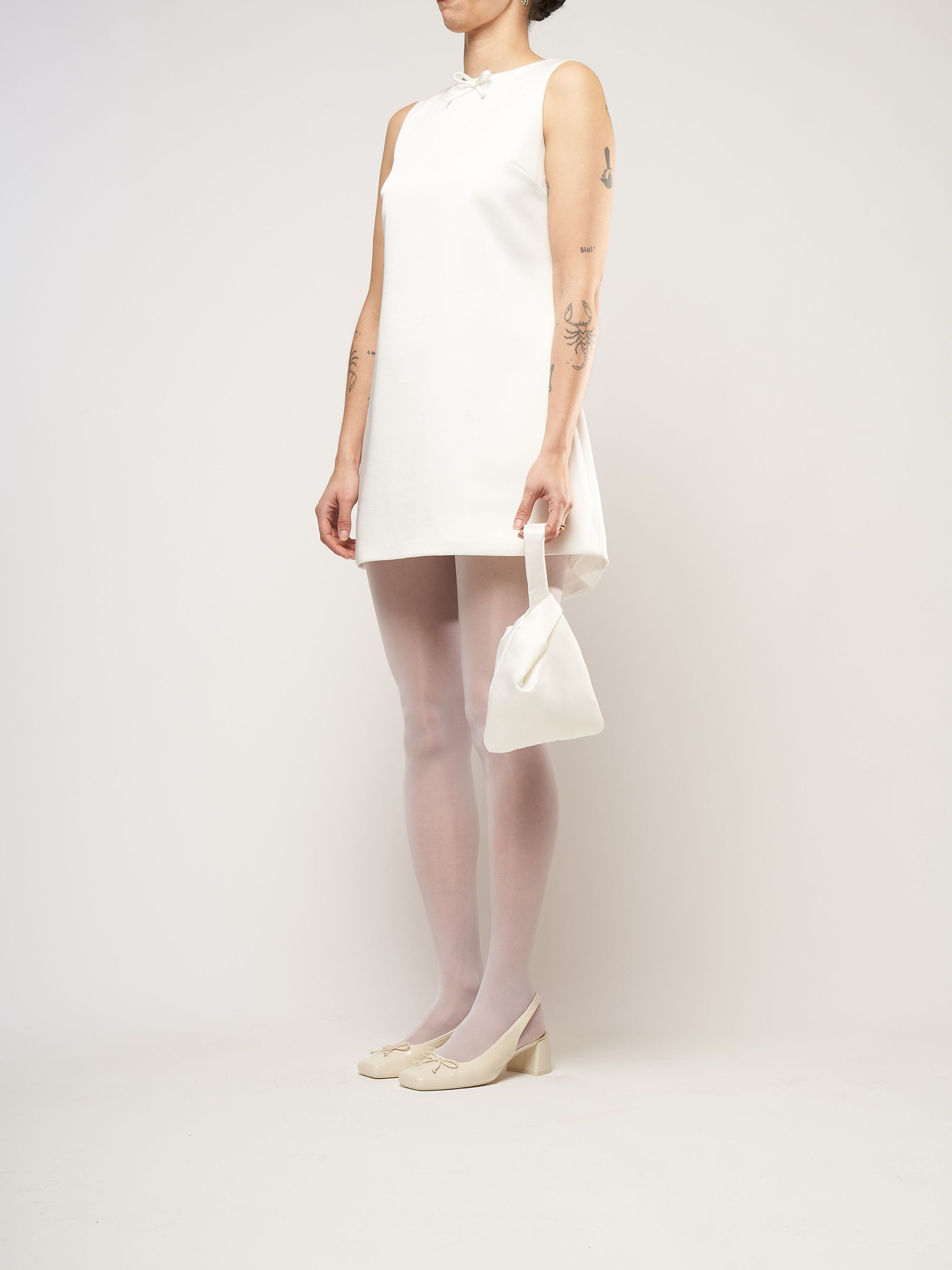 A person in a white sleeveless dress stands against a plain background. They hold a small ivory bag and wear white tights with cream heels.