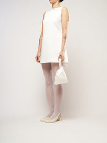 A person in a white sleeveless dress stands against a plain background. They hold a small ivory bag and wear white tights with cream heels.
