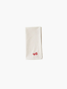 A folded cream-colored cloth napkin with a red embroidered floral design in the corner, set against a plain white background. Simple and elegant.