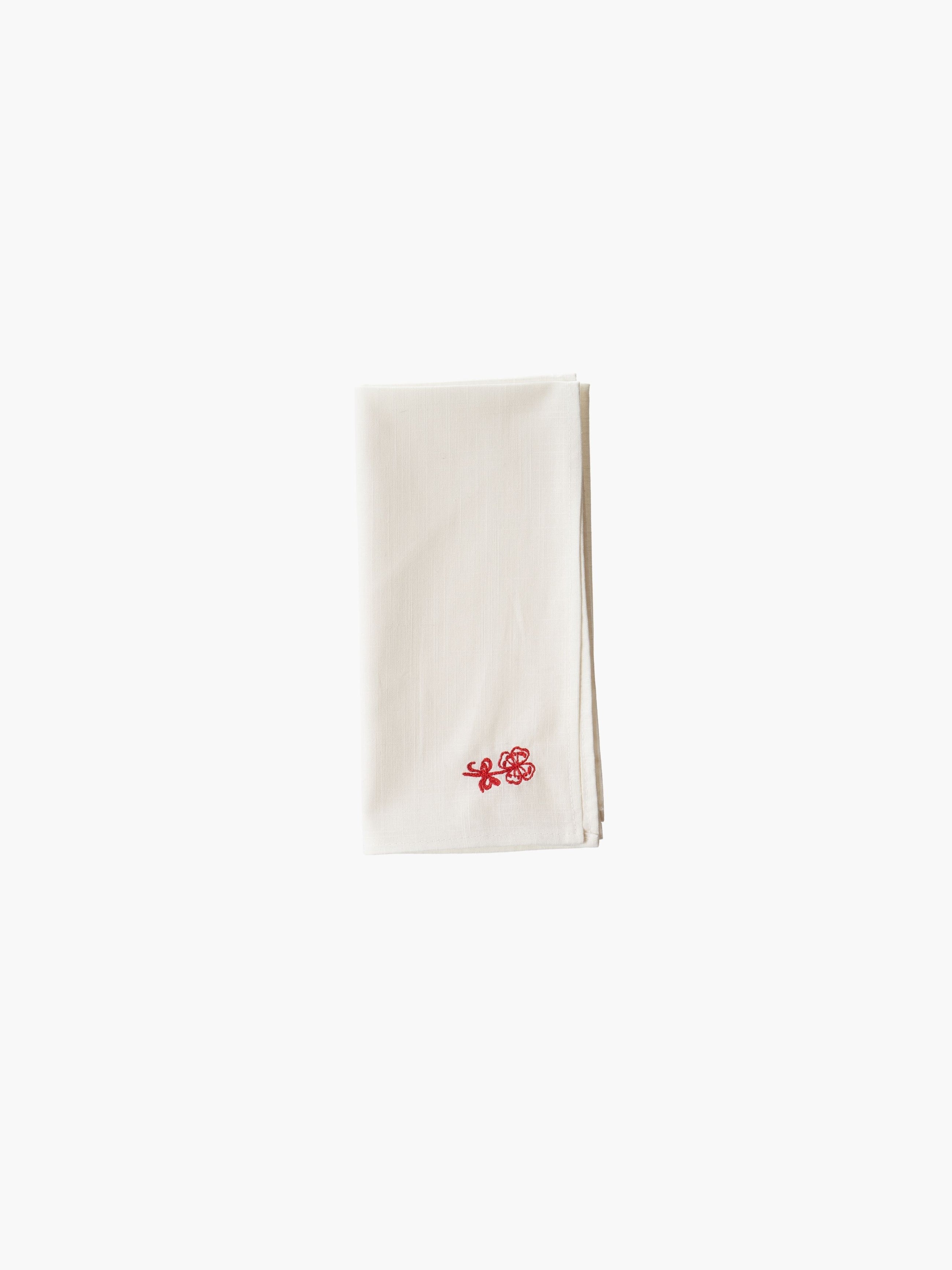 A folded cream-colored cloth napkin with a red embroidered floral design in the corner, set against a plain white background. Simple and elegant.