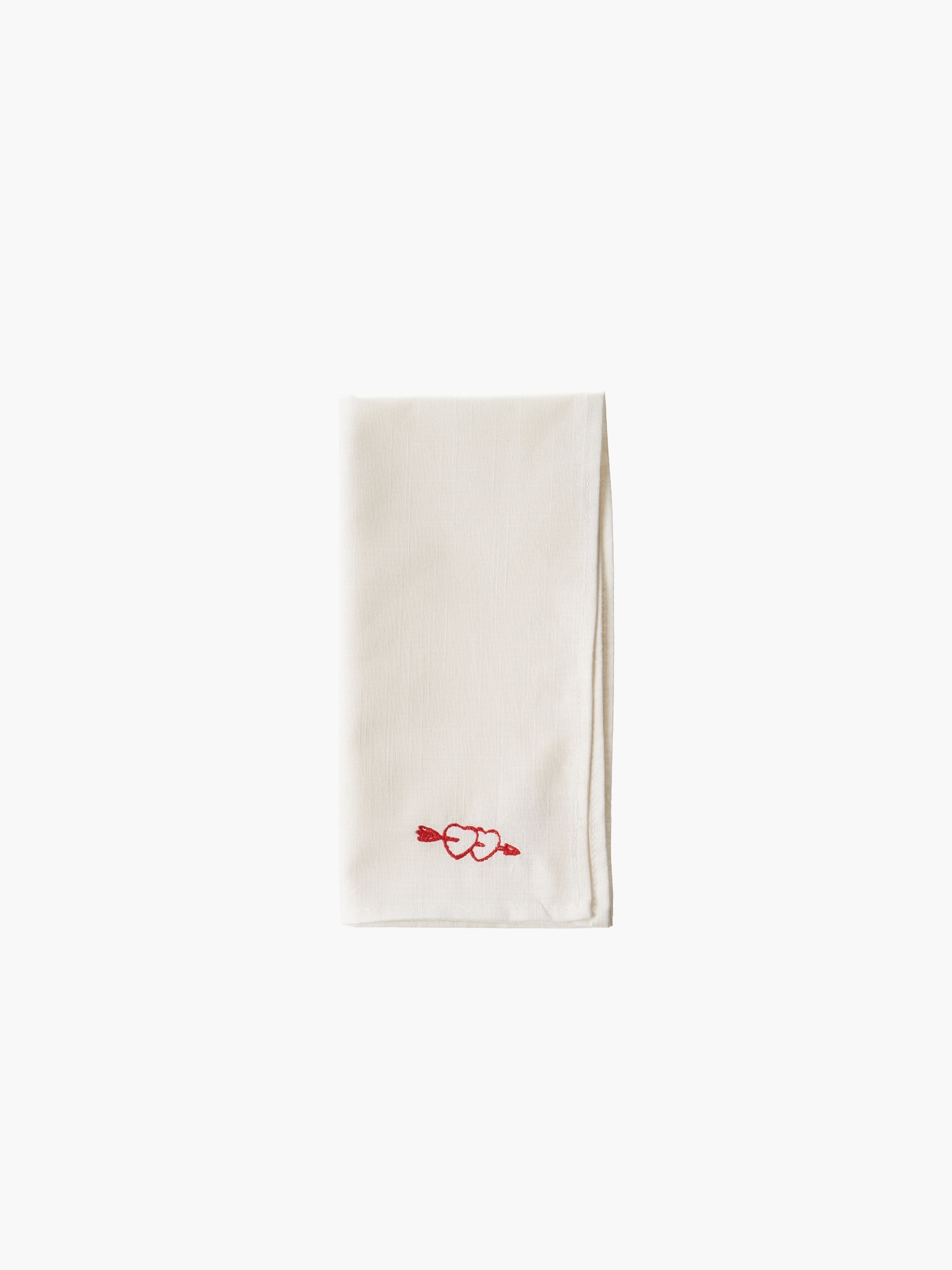 A folded cream-colored cloth napkin with a red embroidered heart design in the corner, set against a plain white background. Simple and elegant.