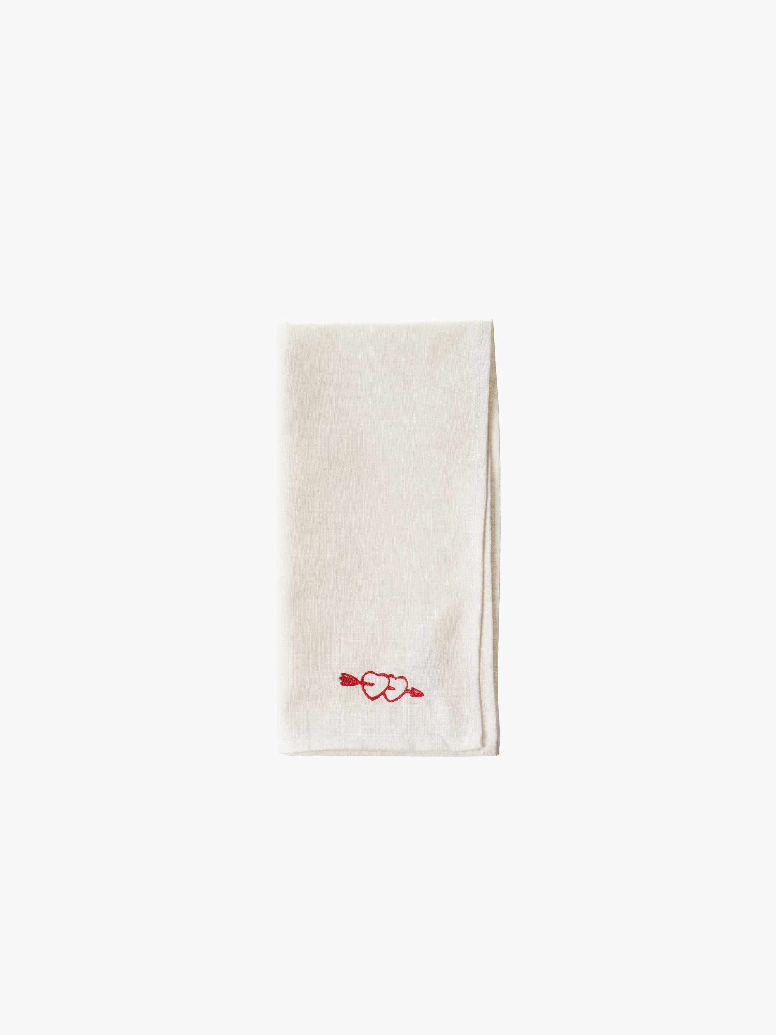 A folded cream-colored cloth napkin with a red embroidered heart design in the corner, set against a plain white background. Simple and elegant.