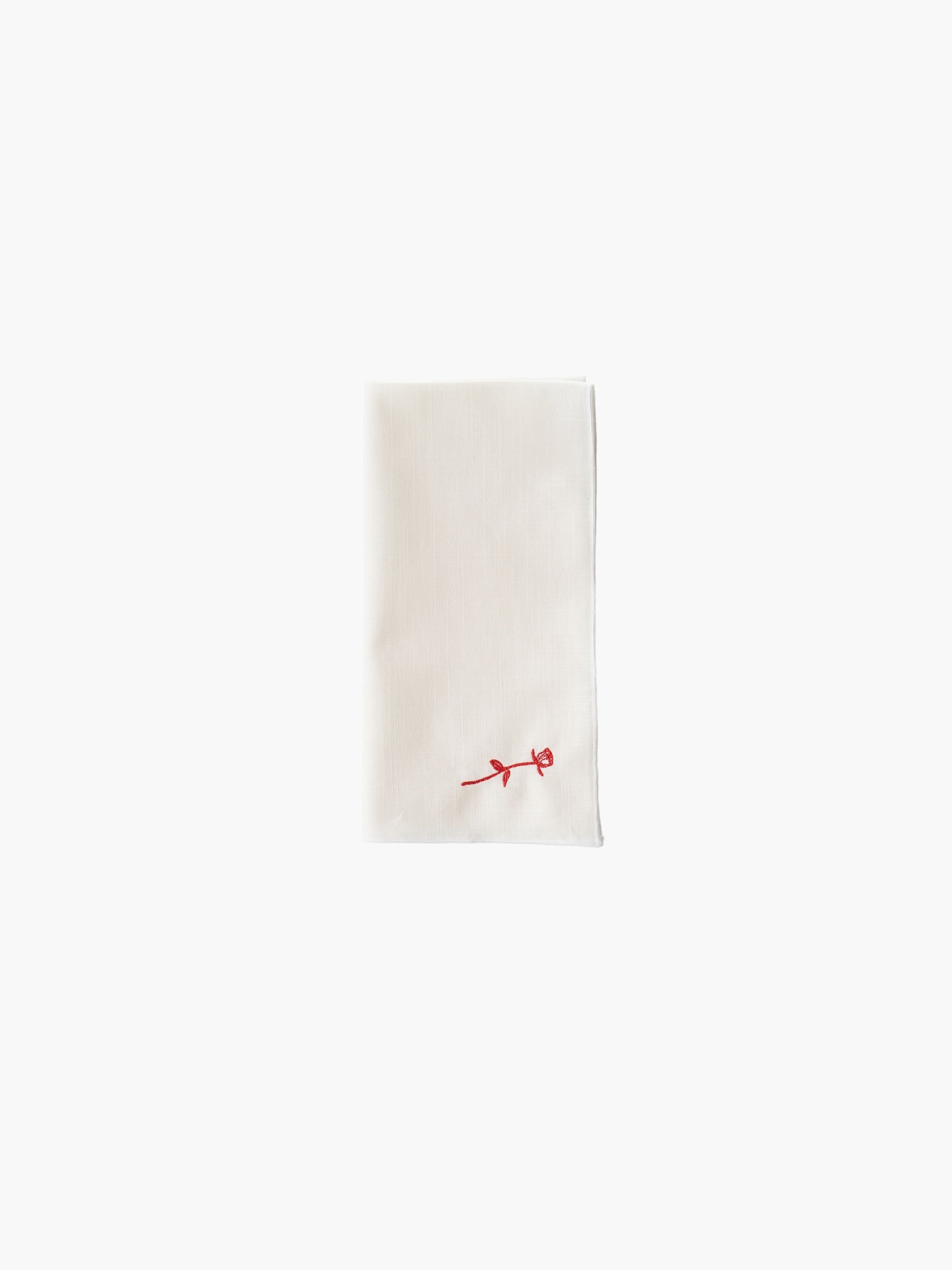 A folded cream-colored cloth napkin with a red embroidered rose design in the corner, set against a plain white background. Simple and elegant.