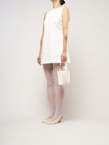 A person in a white sleeveless dress stands against a plain background. They hold a small ivory bag and wear white tights with cream heels.