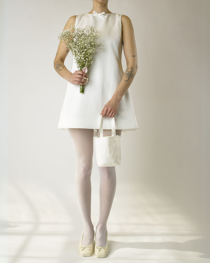 Person wearing an ivory short wedding dress holding a bouquet and a small  ivory satin tote bag against a plain background