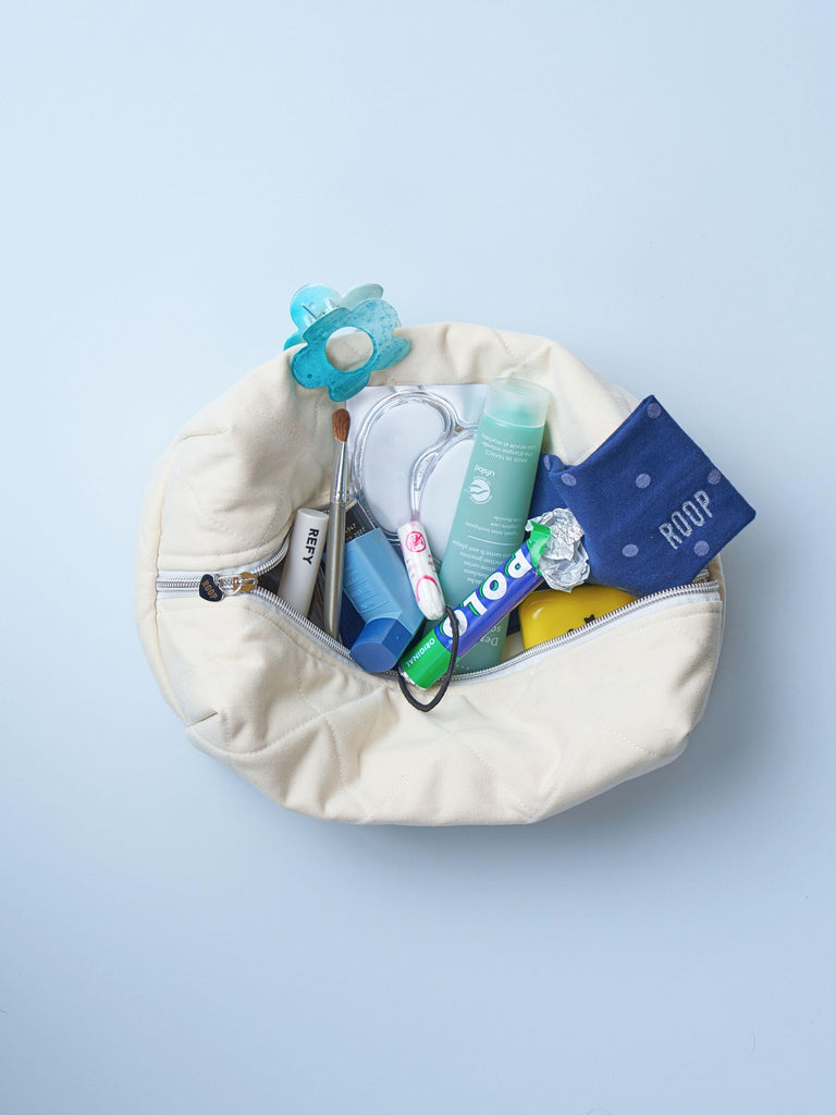 open ivory toiletry bag with cosmetics, pharmaceuticals and hair accessories inside. Set on a pale blue background.