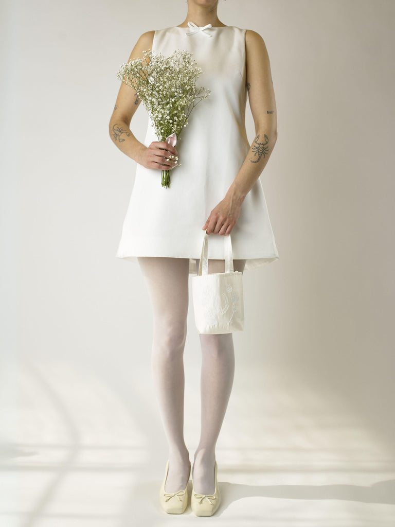 Person wearing a short ivory wedding dress, sheer white tights and ivory shoes, holding a bouquet of flowers and an ivory satin mini tote bag. Set against a plain background. 
