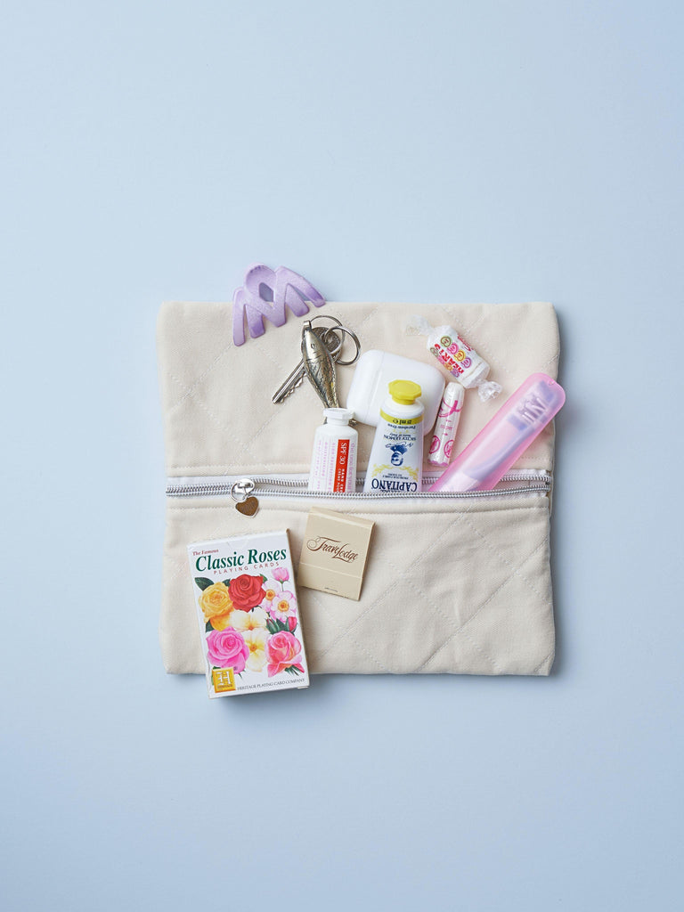 A quilted ivory cotton zipper pouch, the zip has a heart charm on it. Various items are on top of it including a key, toothpaste, and playing cards. Set on a light blue background