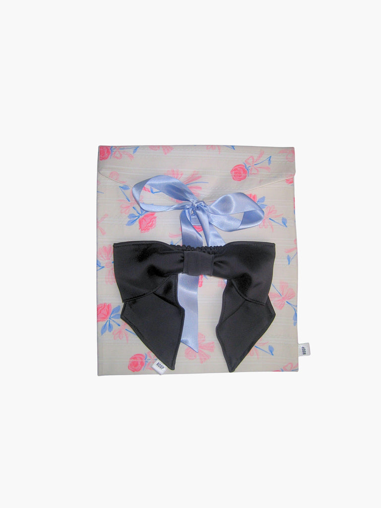 A floral print fabric gift pouch tied with a blue ribbon has a black satin hair bow scrunchie on top of it. Set on a white background.