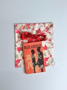 Book titled 'Mom & Me Mom' by Maya Angelou sits on top of a red floral patterned fabric envelope pouch tied with a red ribbon . Set on a pale blue background.