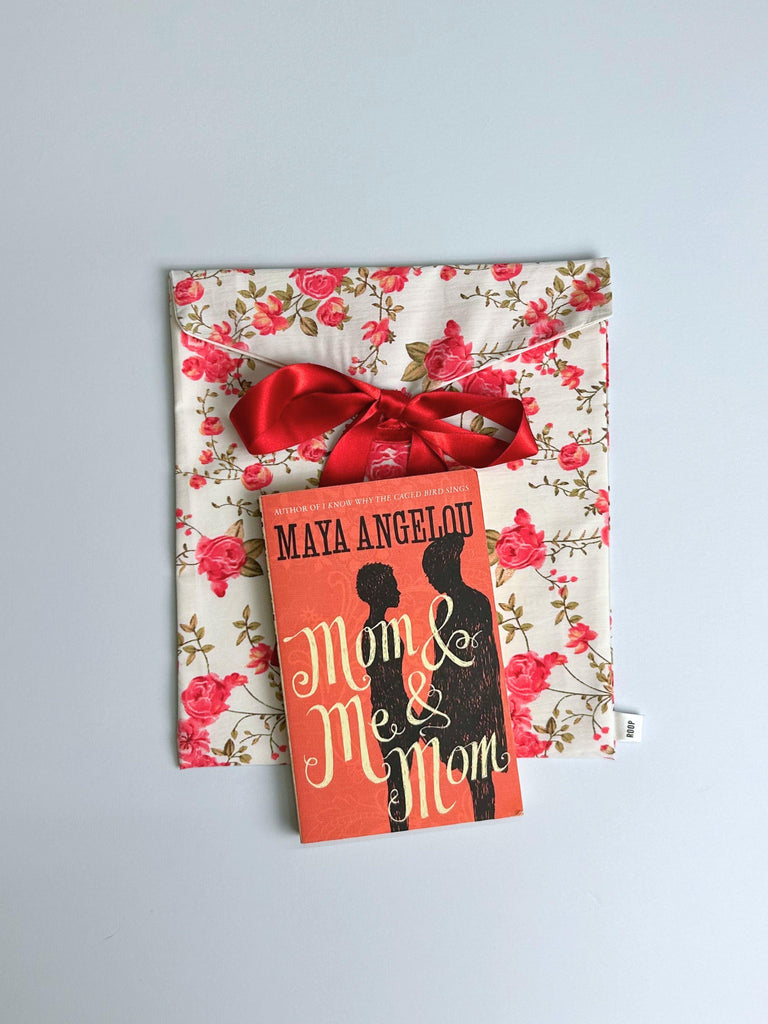 Book titled 'Mom & Me Mom' by Maya Angelou sits on top of a red floral patterned fabric envelope pouch tied with a red ribbon . Set on a pale blue background.