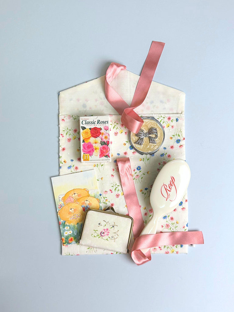 Collection of personal items including a hair brush, playing cards and a compact mirror on top of a floral printed fabric envelope pouch with a pink ribbon. Set on a light blue background.