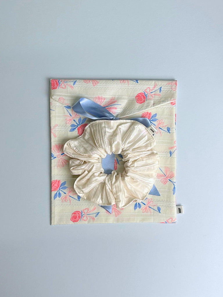 Large ivory taffeta scrunchie sits on top of a floral patterned fabric pouch with a blue  ribbon. Set on a light blue background