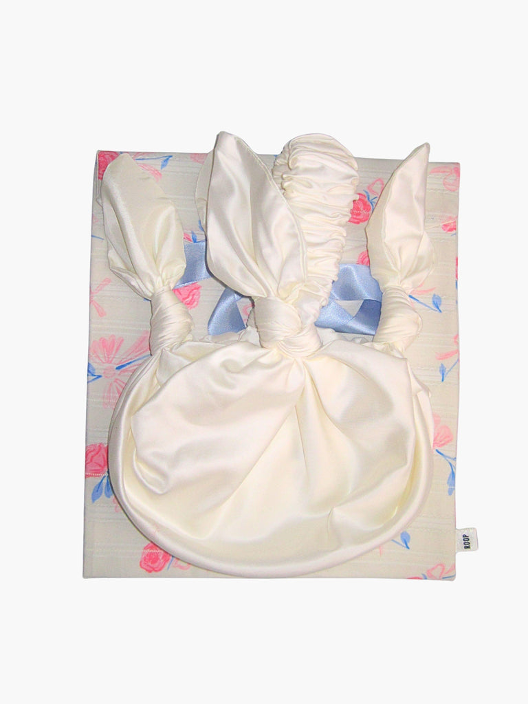 ivory bamboo silk satin knotted handbag, sat on top of a floral patterned ribbon tied fabric git pouch, set against a white background