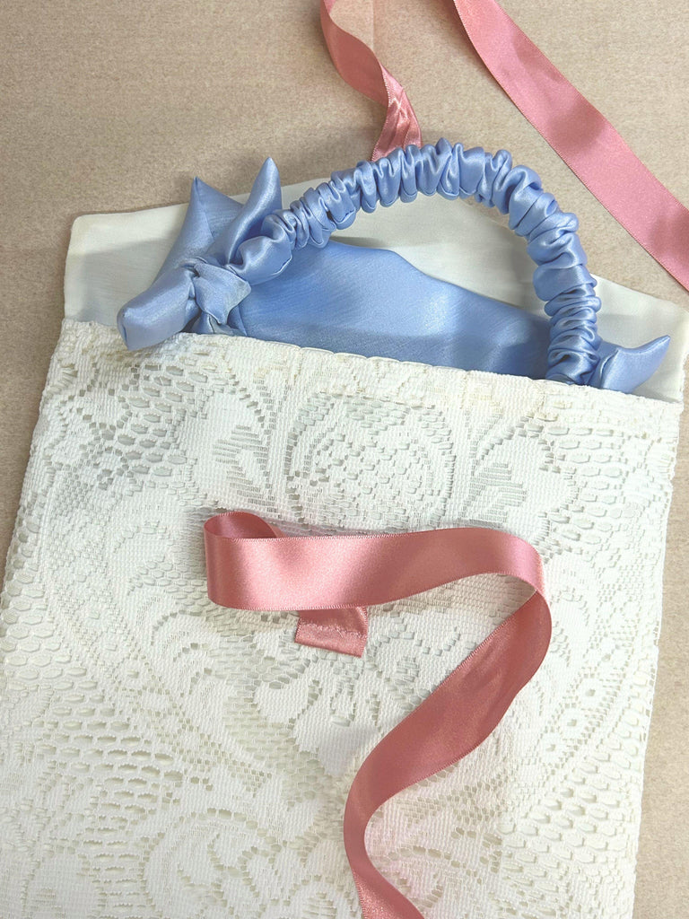 Blue satin handbag with a ruched handle peeks out of a white lace fabric envelope pouch with a pink ribbon. Set on a beige background.