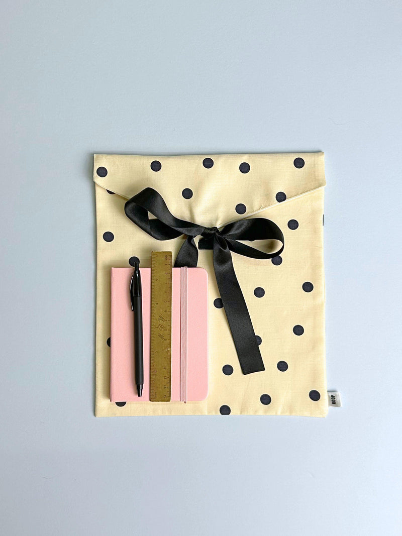 A notebook, ruler and pen sit on top of a cream and black with polka dot patterned fabric envelope pouch tied with a black ribbon. Set on a pale blue background.