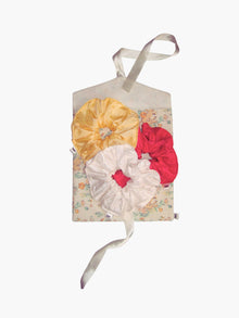 A trio of large scrunchies in yellow, ivory and pink sit on a top of a floral and rabbit print fabric envelope gift pouch tied with an ivory ribbon. Set on a white background