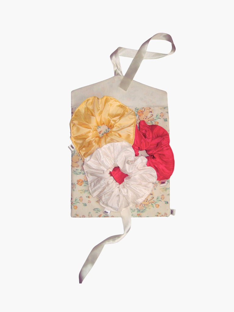 A trio of large scrunchies in yellow, ivory and pink sit on a top of a floral and rabbit print fabric envelope gift pouch tied with an ivory ribbon. Set on a white background