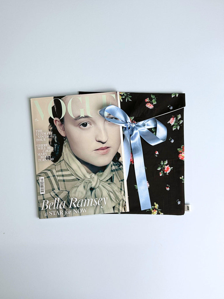 Vogue magazine next to a black floral envelope pouch  tied with a blue ribbon, on a light blue background