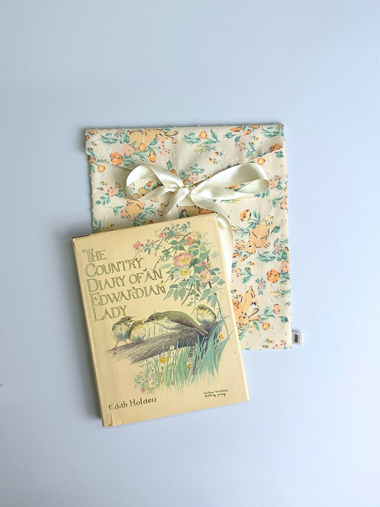 Book titled 'The Country Diary of an Edwardian Lady' with a decorative cover, sits on top of a floral and rabbit print fabric envelope pouch tied with an ivory ribbon. Set on a light blue background.
