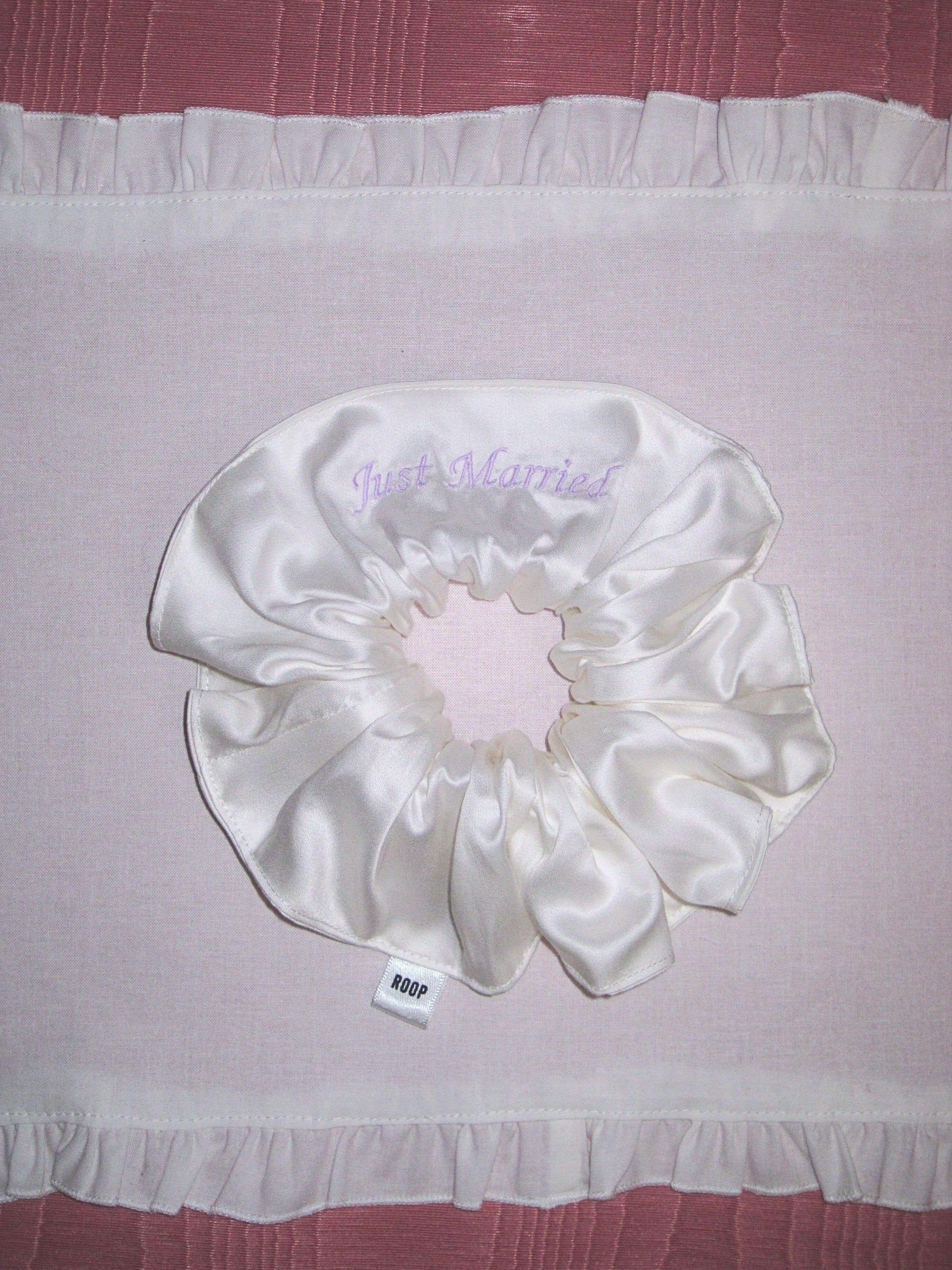 Ivory bamboo silk satin scrunchie with 'Just Married' embroidered in lilac on a white ruffled placemat, set on a pink moire  background.