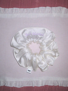 Ivory bamboo silk satin scrunchie with 'Just Married' embroidered in lilac on a white ruffled placemat, set on a pink moire  background.
