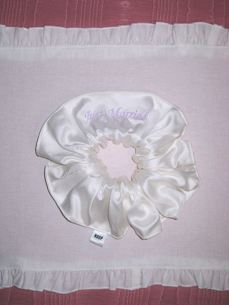 Ivory bamboo silk satin scrunchie with 'Just Married' embroidered in lilac on a white ruffled placemat, set on a pink moire  background.