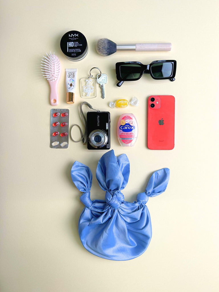 Collection of everyday items including a phone, camera, and makeup products on a beige background. A lilac handbag sits at the bottom.