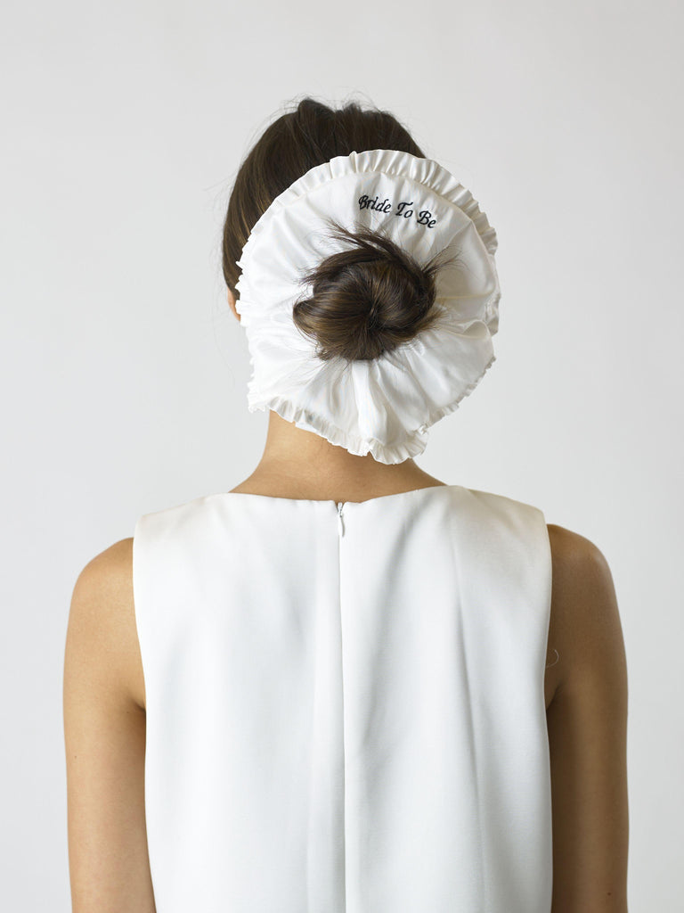 Person wearing an Ivory ruffled scrunchie over their bun with black embroidery that reads 'bride to be', set on a white background