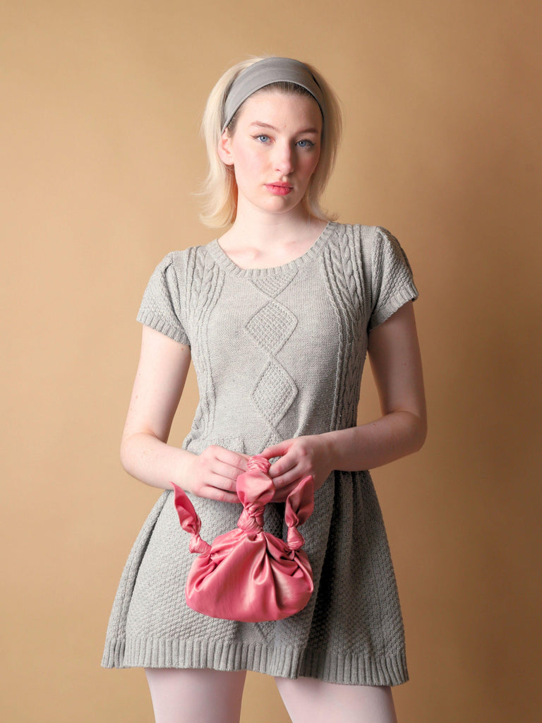 Woman wearing a gray knit dress holding a pink knotted bag against a beige background