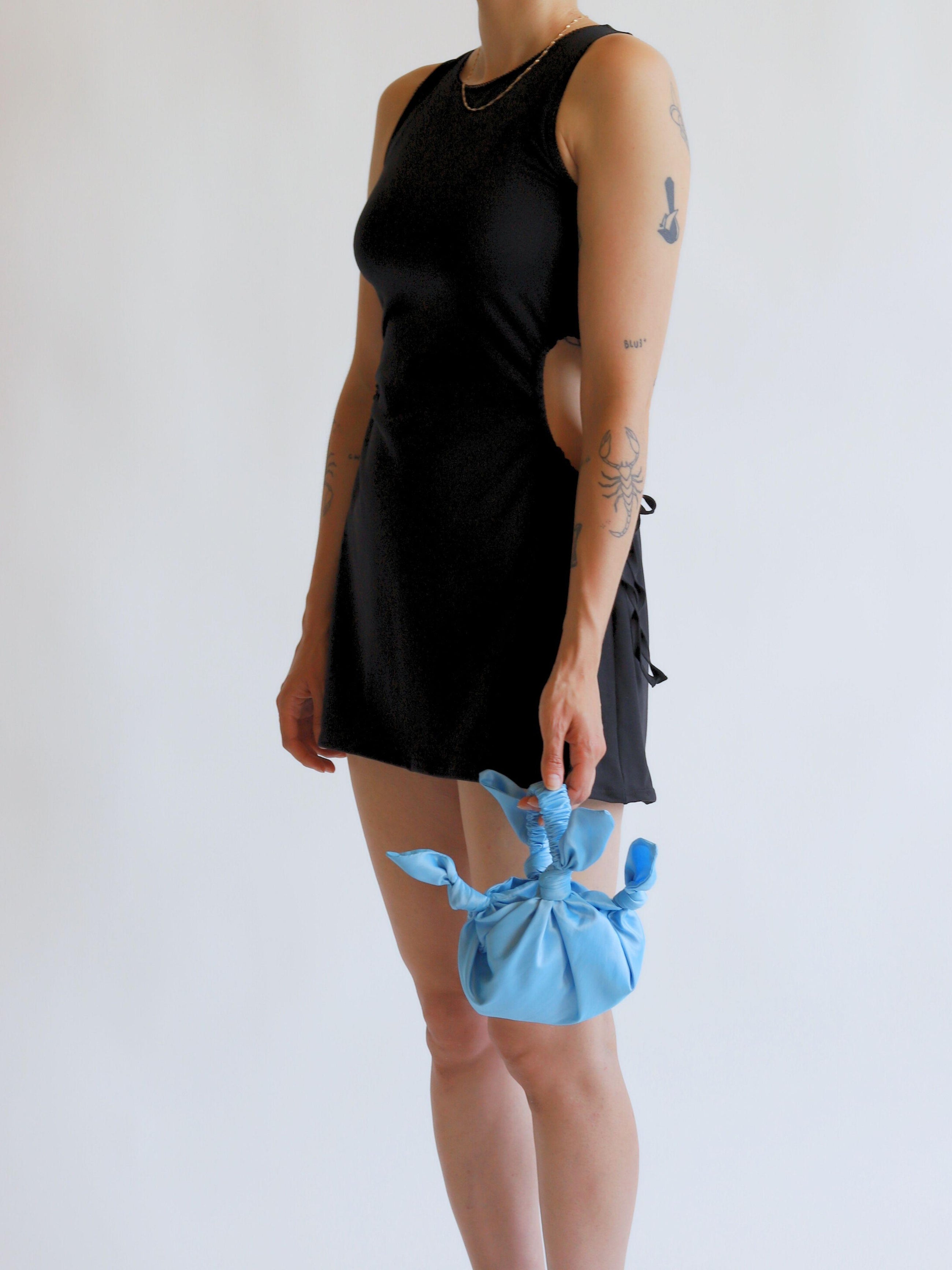 Person wearing a black sleeveless dress holding a blue knotted bag against a white background