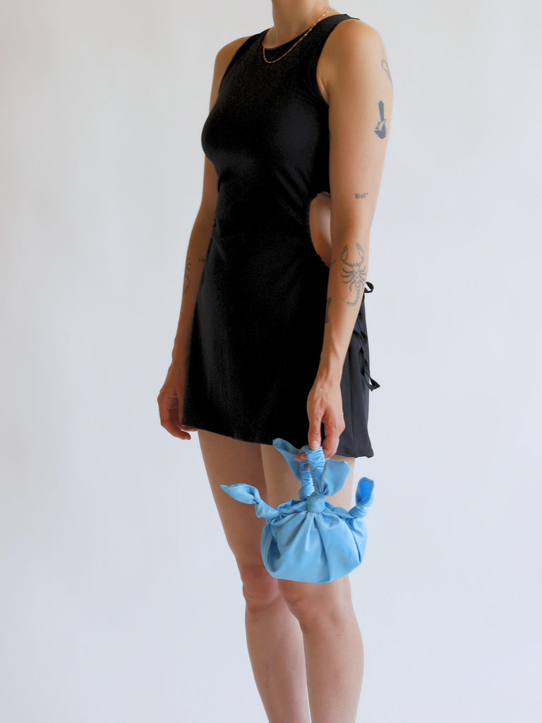 Person wearing a black sleeveless dress holding a blue knotted bag against a white background