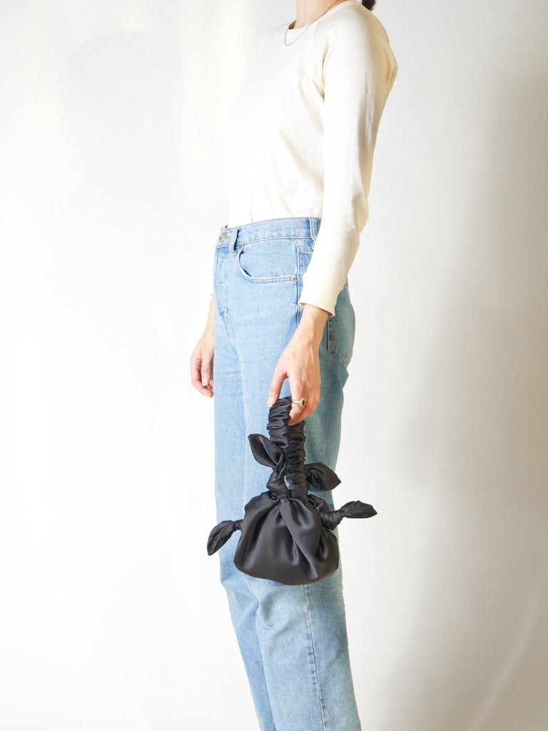 Person wearing blue jeans holding a black satin knotted bag against a plain background