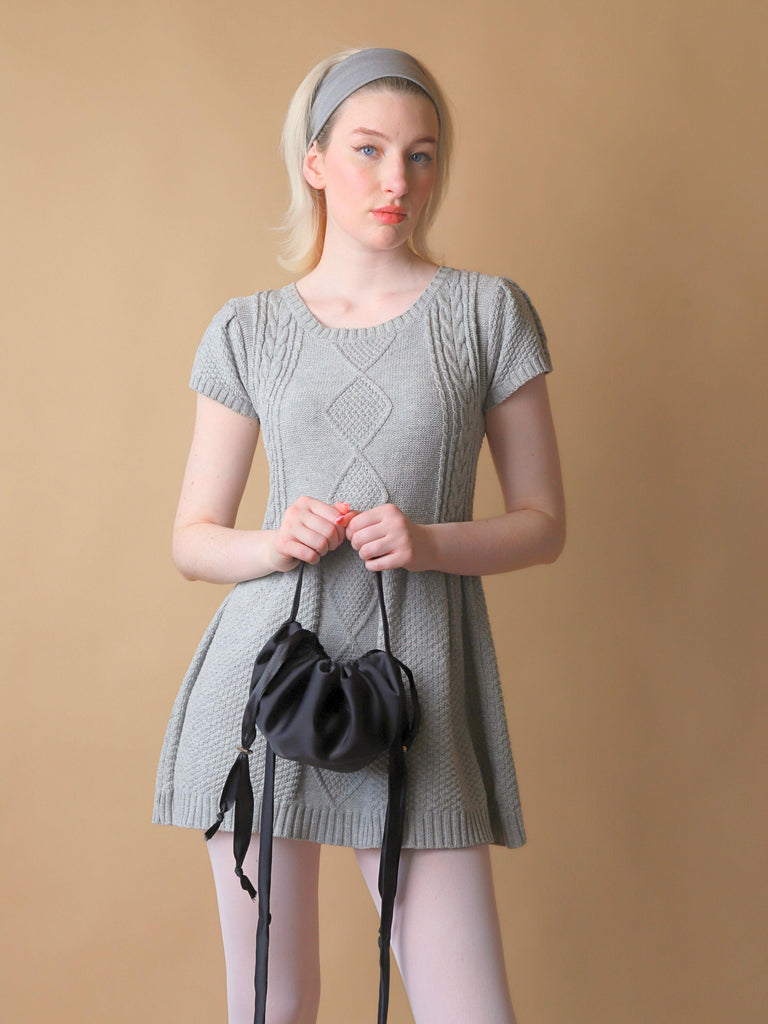 Woman wearing a grey knit dress holding a black satin puffball drawstring bag in front of her, set against a beige background
