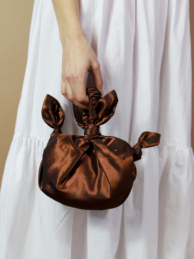 Brown taffeta knotted handbag held by a person wearing a white dress against a neutral background
