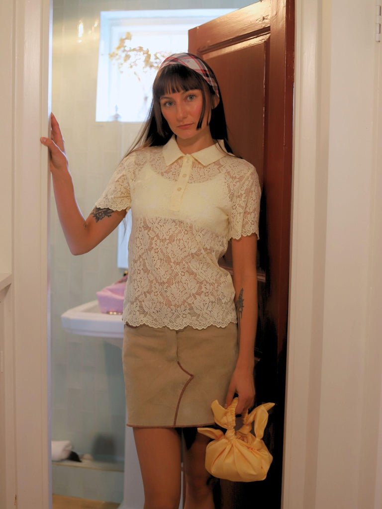 Woman wearing a lace top and beige skirt in a bathroom setting, she holds a yellow knotted handbag by her side.