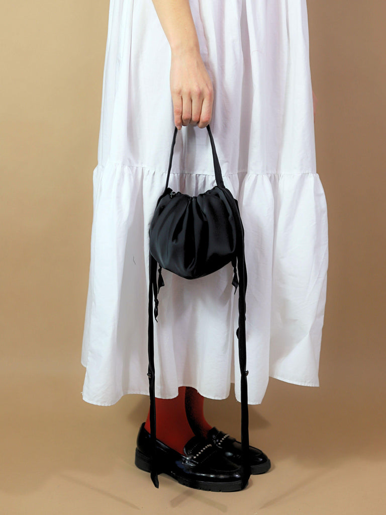 Person wearing a white tired maxi dress, red tights and black loafers, holds a black satin puffball drawstring handbag, set on a beige background.