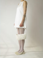 Person wearing a short ivory wedding, sheer white stocking and heeled ivory ballet pumps, holds an ivory satin textured ruched front handbag. Set on a white background.