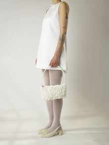 Person wearing a short ivory wedding, sheer white stocking and heeled ivory ballet pumps, holds an ivory satin textured ruched front handbag. Set on a white background.