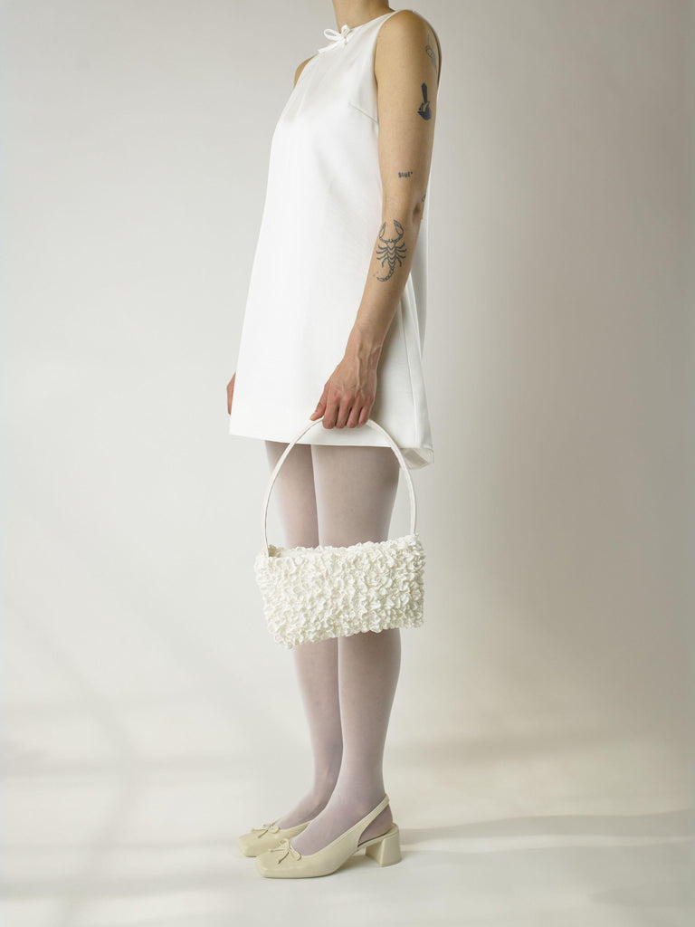 Person wearing a short ivory wedding, sheer white stocking and heeled ivory ballet pumps, holds an ivory satin textured ruched front handbag. Set on a white background.