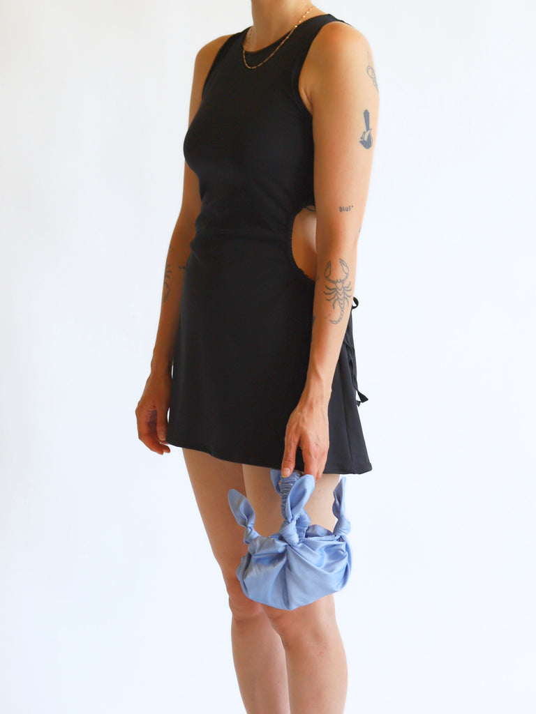 Person wearing a black sleeveless dress holding a lilac knotted handbag on a white background