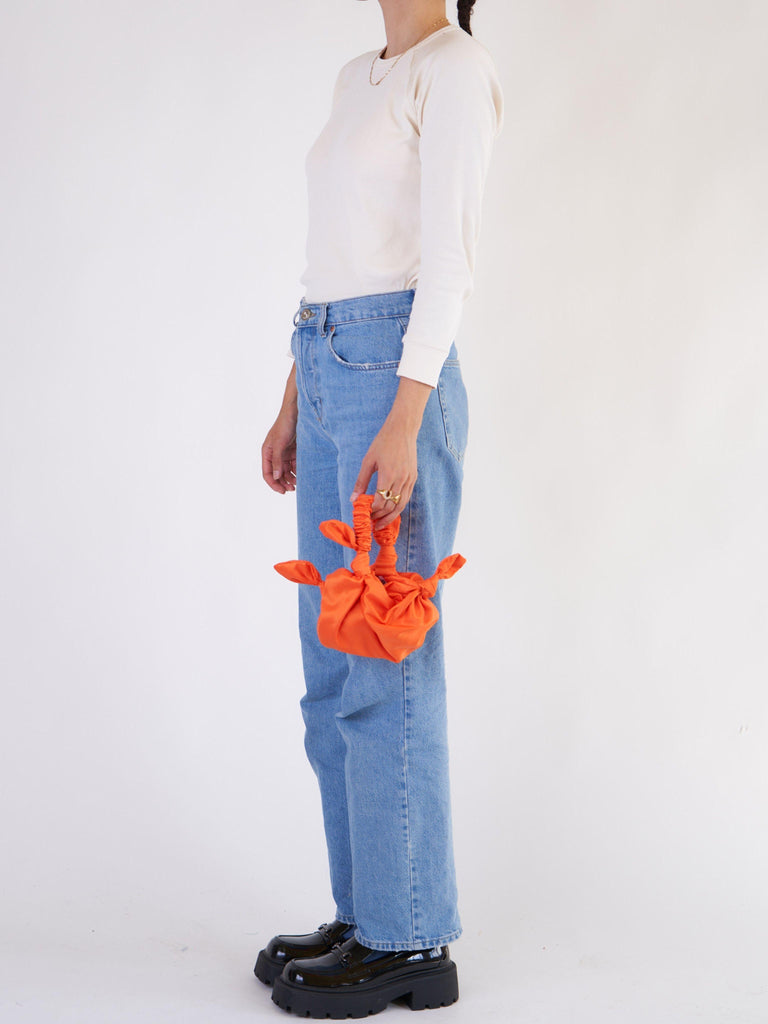 Person wearing blue jeans and a white top, holding an orange knotted handbag against a white background