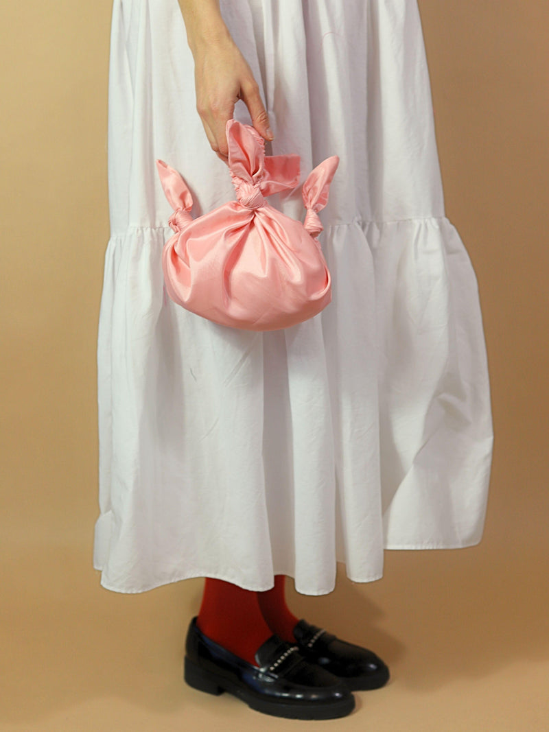 Person holding a pink knotted bag wearing a white dress, red tights and black loafers against a beige background