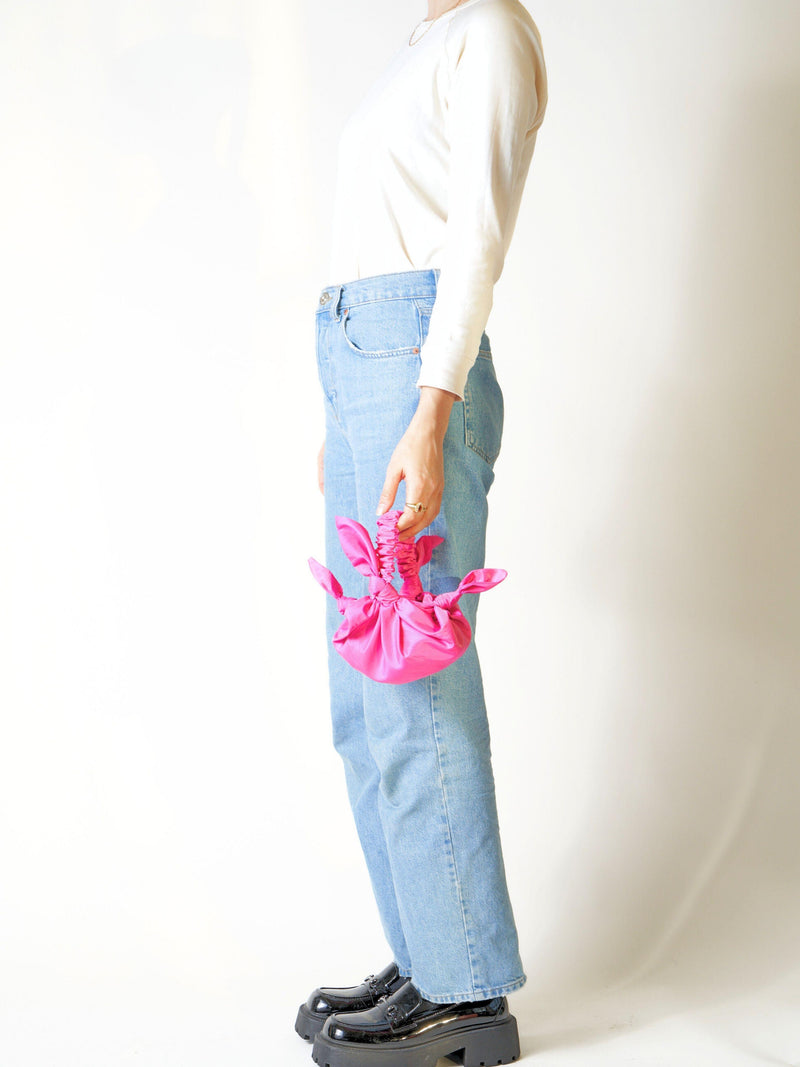 Person wearing light blue jeans holds a pink knotted bag held in hand against a white background