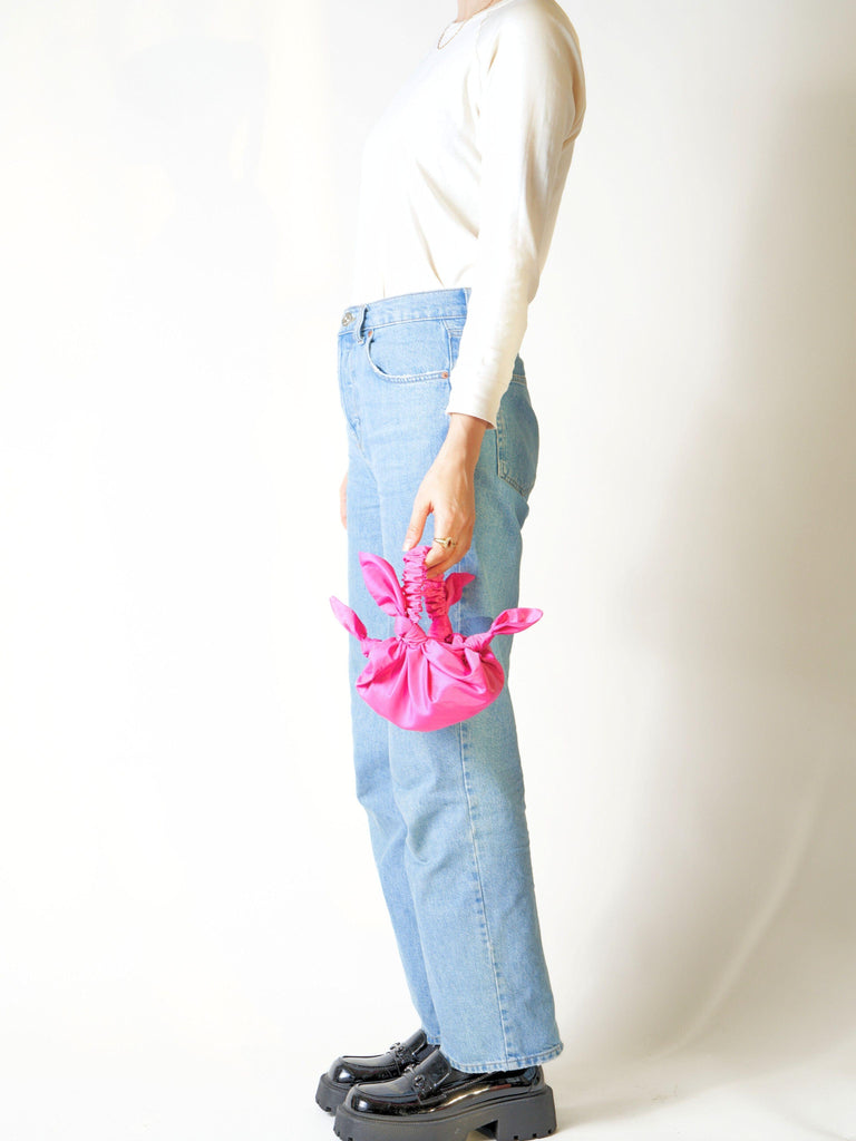 Person wearing light blue jeans holds a pink knotted bag held in hand against a white background