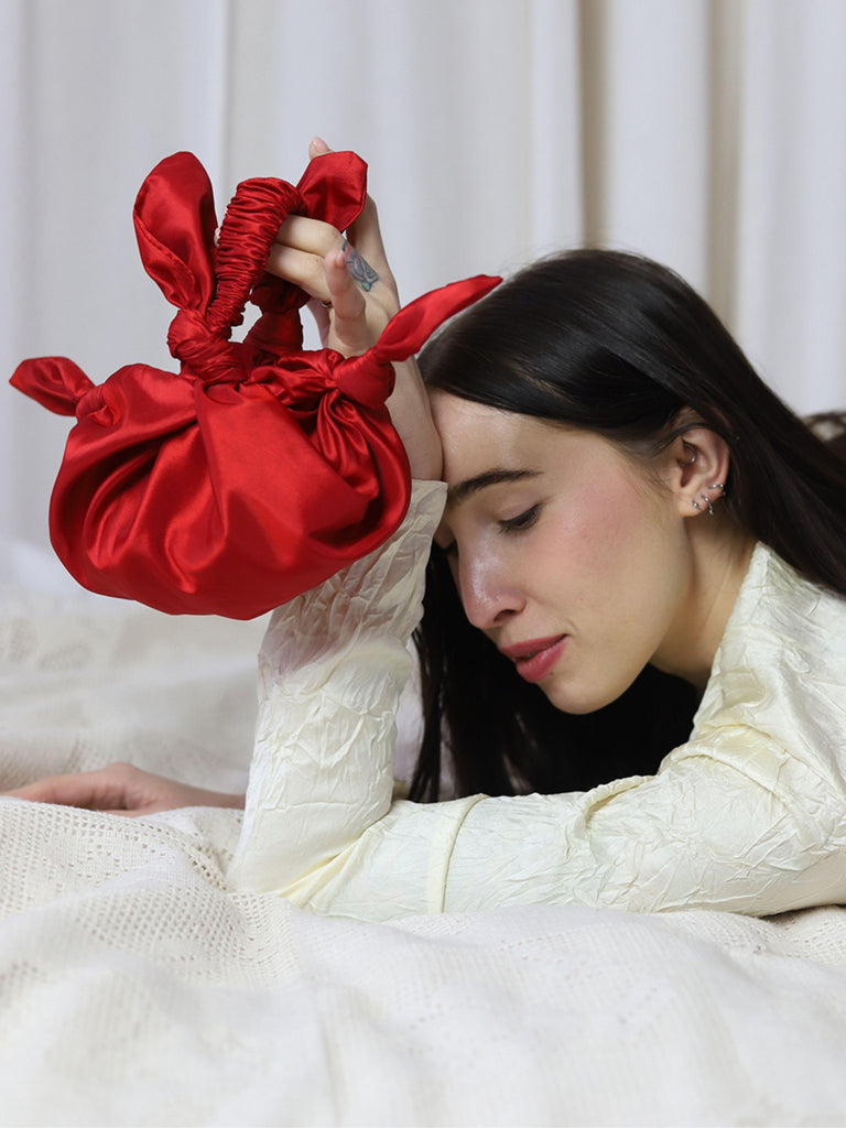 Woman holding a red taffeta knotted bag against a white curtain background