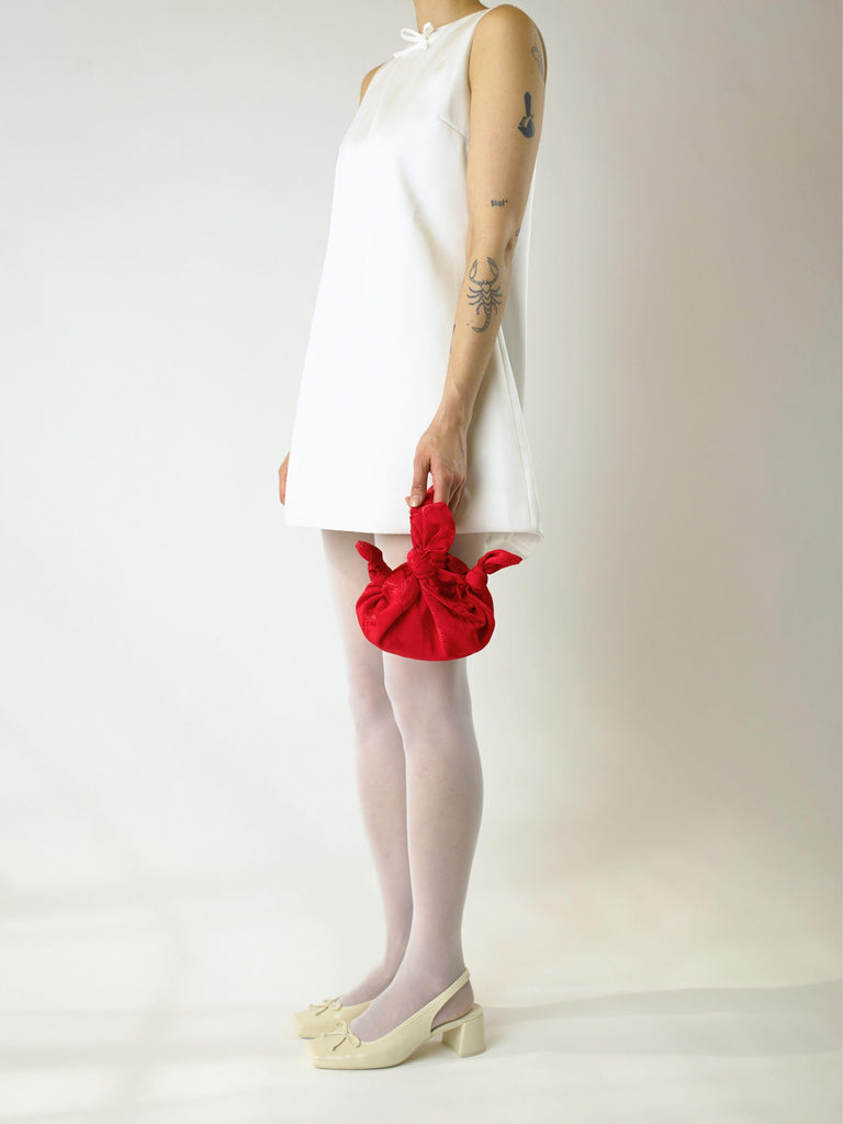 Person in an ivory short wedding dress holding a red knotted floral satin handbag against a plain background